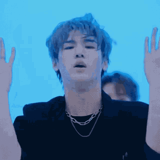 a young man wearing a black jacket and a necklace is standing in front of a blue background .