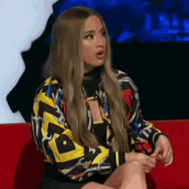 a woman is sitting on a red couch wearing a colorful jacket and a black skirt .