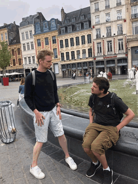 two men standing and sitting in front of a fountain in front of a building that has a sign that says ' boutique '