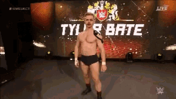 a shirtless wrestler is standing on a stage in front of a large screen that says tyler bate .