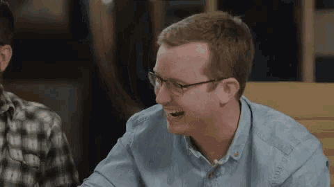 a man wearing glasses is laughing while sitting at a table with another man .