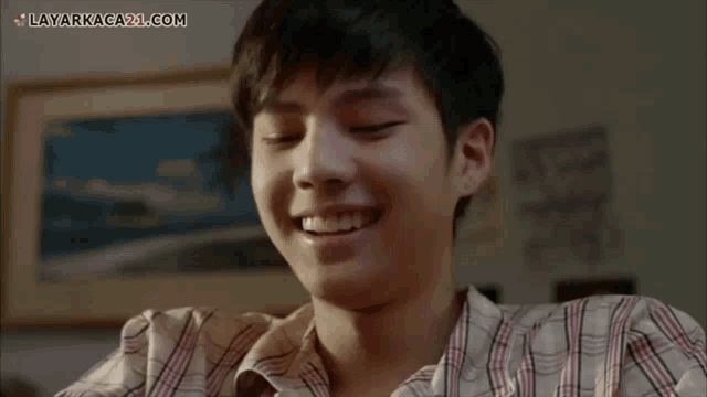a young man in a plaid shirt is smiling with the website layarkaca21.com visible in the corner