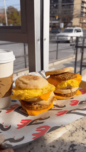 two breakfast sandwiches are on a tray with a cup of coffee in the background