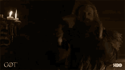 a man with a beard is holding a sword in front of two lit candles and the word got is on the bottom