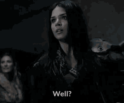 a woman in a black jacket is standing in a dark room and talking to someone .