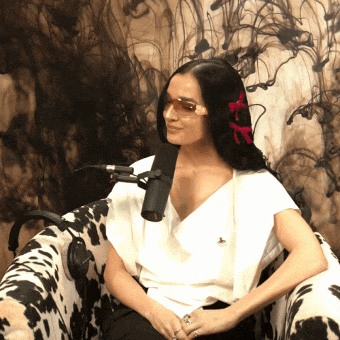a woman wearing sunglasses sits in a chair with a microphone in front of her
