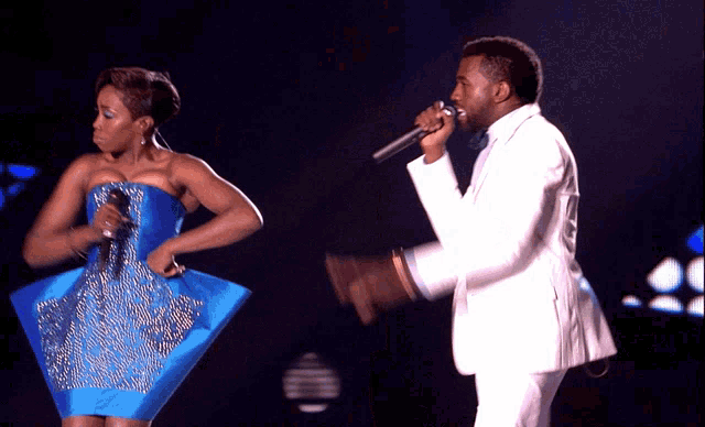a man in a white suit is singing into a microphone while a woman in a blue dress stands behind him