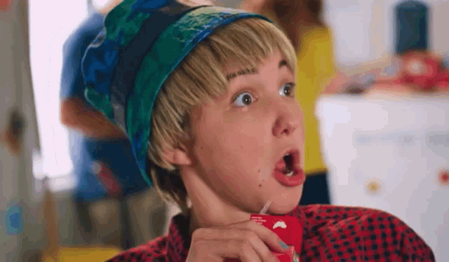 a young man wearing a beanie and a plaid shirt is drinking from a red can with a straw .