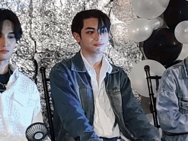 a man in a denim jacket is sitting in front of balloons and a fan .