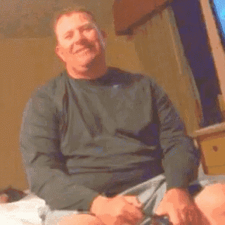a man in a black shirt is sitting on a bed holding a remote control .