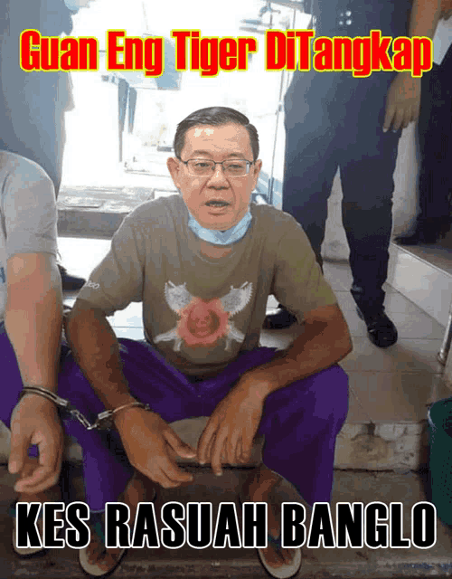 a man in handcuffs is sitting in front of a sign that says guan eng tiger di angkap kes rasuah banglo
