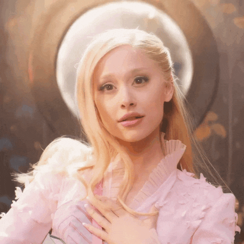 a blonde woman in a pink dress holds her hands to her chest