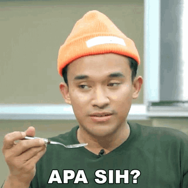 a man wearing an orange beanie is holding a spoon and has the word apa sih on his shirt