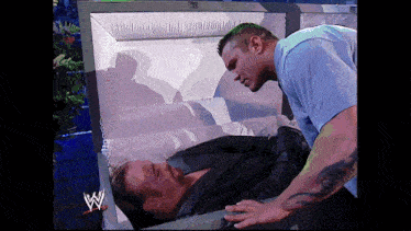 a man in a blue shirt is standing next to a coffin with a w logo on it
