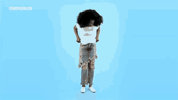 a woman wearing ripped jeans and a white t-shirt stands in front of a blue background with the website cosmopolitan.com visible