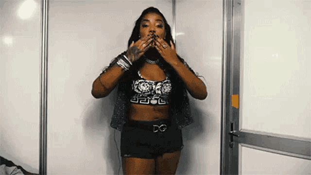 a woman in a crop top and shorts is standing in front of a white wall and covering her mouth with her hands .