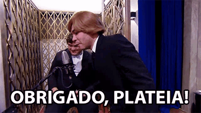 a man in a suit and tie is standing next to another man in front of a microphone and says obrigado plateia .