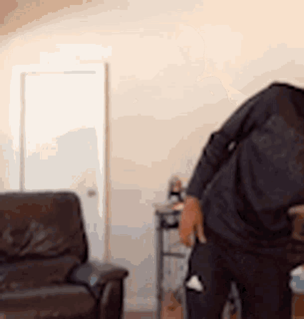 a man in a black shirt is standing in a living room next to a chair .
