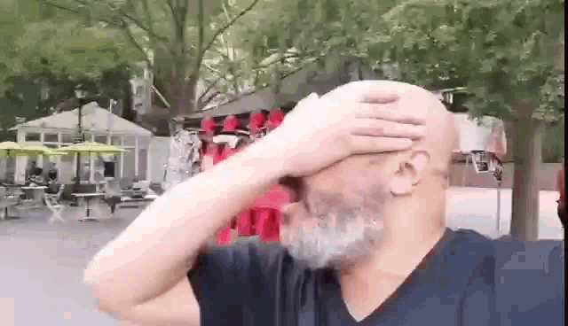 a bald man with a beard is covering his face with his hand in a park .