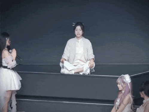 a woman in a white dress sits on a ledge with her legs crossed while another woman sings into a microphone