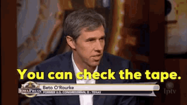 a man in a suit is sitting in front of a sign that says you can check the tape .