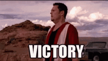 a man in a red and white vest is standing in the desert with the words `` victory '' above him .
