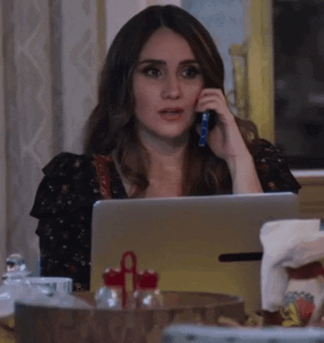 a woman is sitting at a table talking on a cell phone while using a laptop .