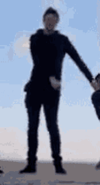 a man in a black shirt is standing on a sand dune holding another man 's hand .