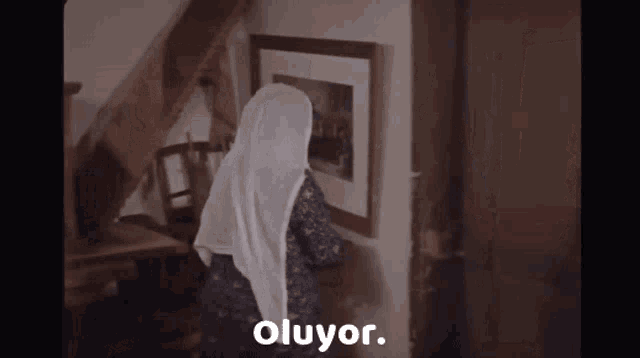 a woman in a white head scarf is looking at a picture on a wall and the word oluyor is visible