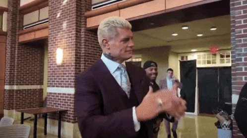 a man in a suit clapping his hands in front of a brick wall