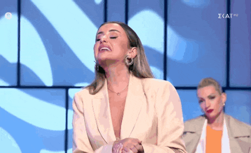 a woman in a tan jacket is laughing while another woman stands behind her with the letters kat on the bottom