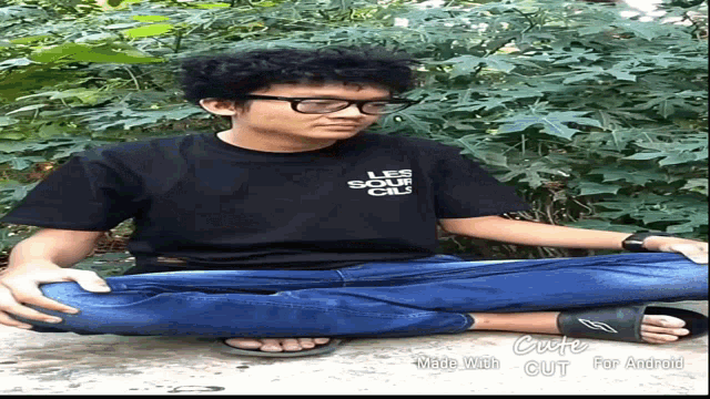 a man wearing glasses and a black shirt that says les sour cils sits on the ground