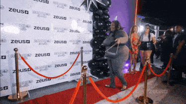 a woman is standing on a red carpet in front of a wall that says zeus baddies