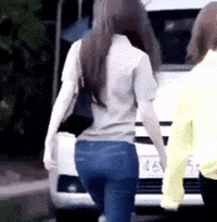 a woman in a gray shirt and blue jeans is walking down the street next to a car .