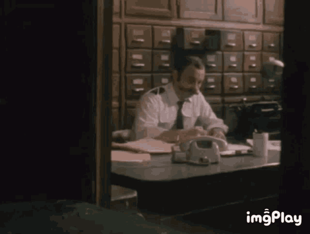 a man with a mustache sits at a desk in a dark room with images play written on the bottom