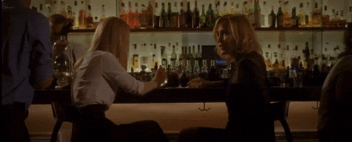 two women are sitting at a bar talking to each other with bottles of alcohol behind them