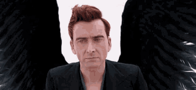 a man with red hair and black wings is sitting in front of a white wall .