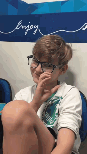 a boy wearing glasses is sitting in front of a wall with the word enjoy written on it