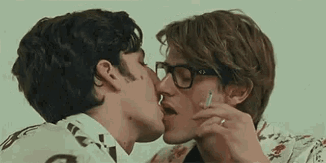 two men are kissing and smoking a cigarette .