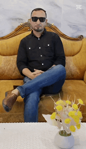 a man wearing sunglasses sits on a couch next to a vase of yellow flowers