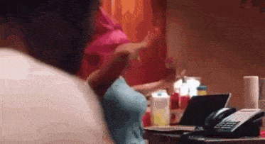 a woman in a pink shirt is sitting at a table with a phone and a laptop .