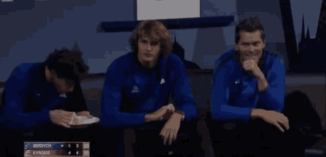three men in blue jackets sit on a couch with a scoreboard behind them that says berdych