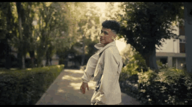 a young man in a white jacket is walking down a sidewalk in a park