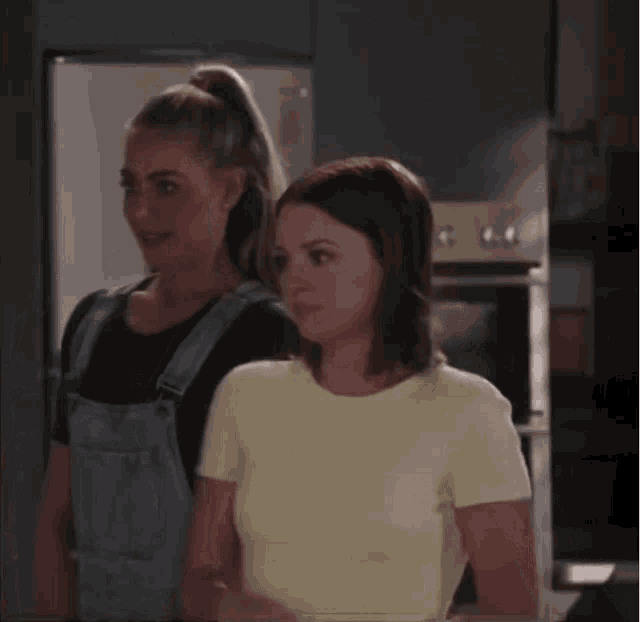 two women standing next to each other with one wearing overalls and the other a yellow shirt