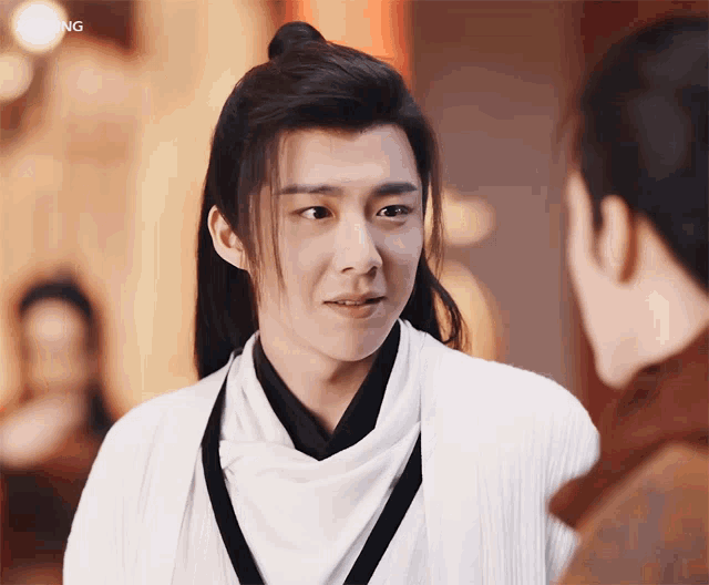 a man with long hair and a ponytail looks at another man in front of a blurred background with the word qing visible
