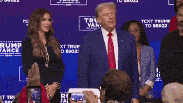 a man in a blue suit and red tie stands in front of a sign that says trump vance