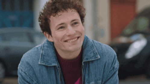 a man with curly hair wearing a denim jacket smiles