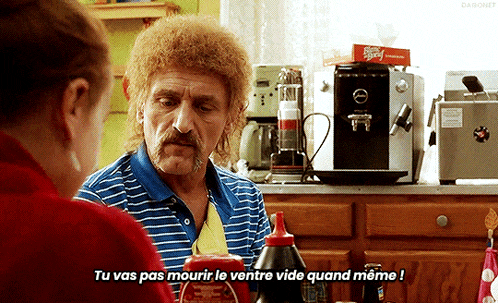 a man with a mullet is talking to a woman in a kitchen with a ketchup bottle in front of him