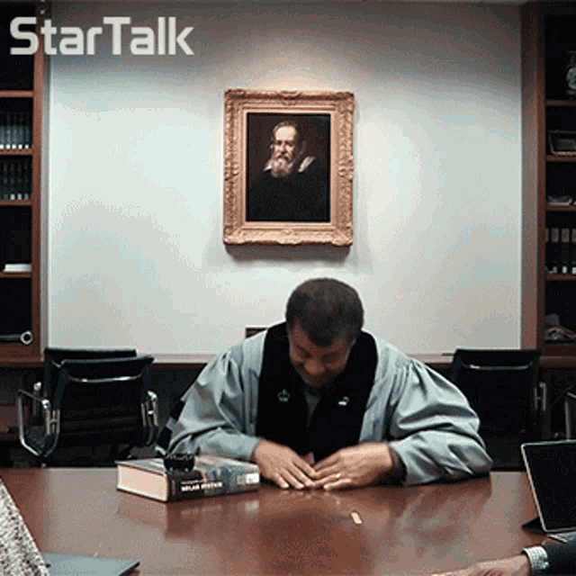 a man sits at a table in front of a framed portrait of a man with a beard and the words startalk above him