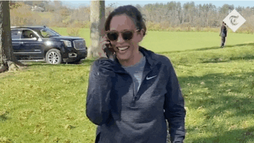 a woman wearing sunglasses is talking on a cell phone while standing in a field .